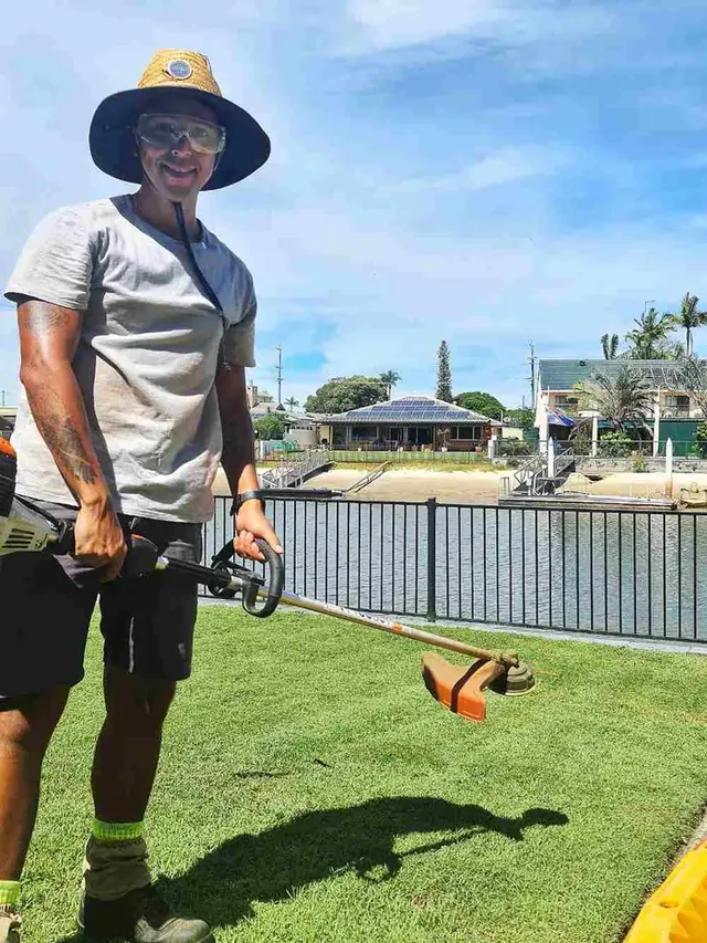 Regis, a Your Helpers gardener in a wide-brim hat, holding a whipper snipper on a canal-side lawn
